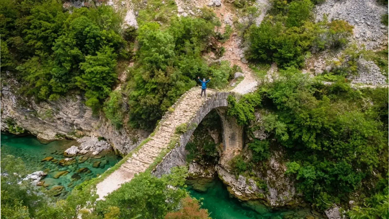 Hiker standing on a humped stone bridge above turquoise water in a green gorge