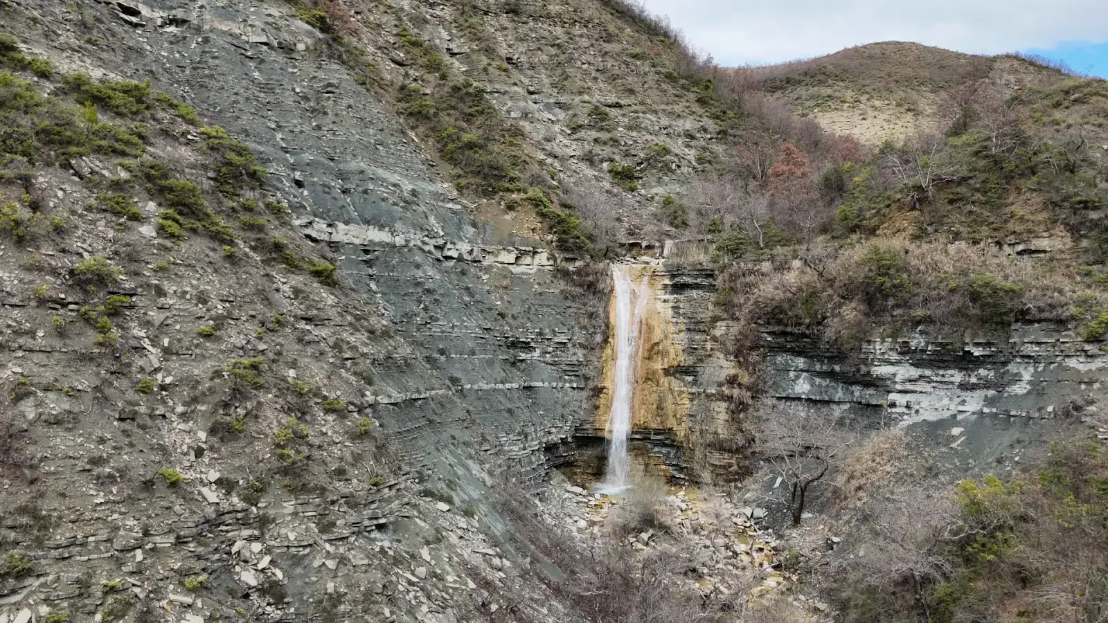 Vista aérea de una alta cascada que cae por acantilados grises estratificados cerca de Podë