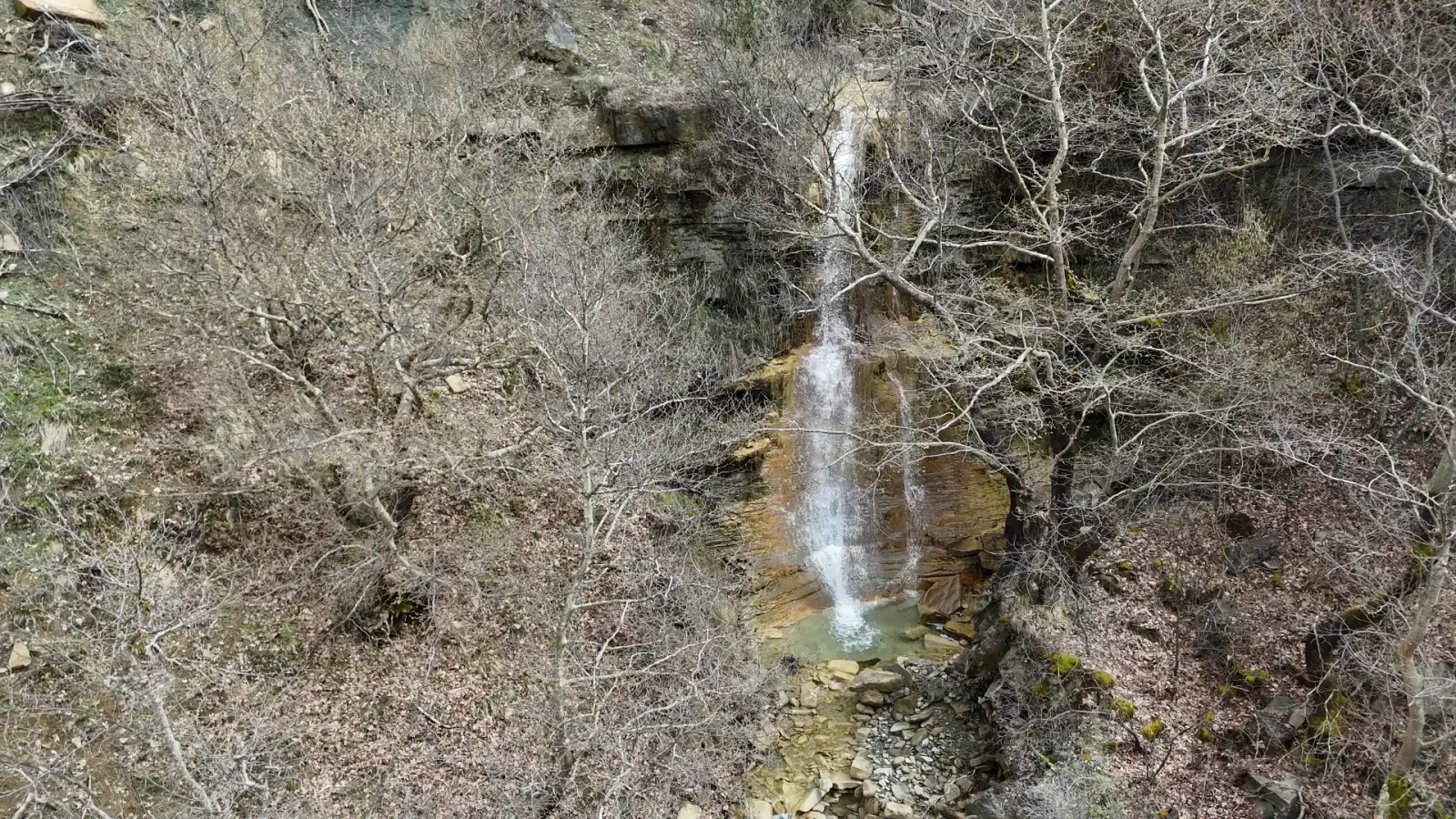 Esbelta cascada en Podë entre árboles desnudos invernales en una ladera rocosa