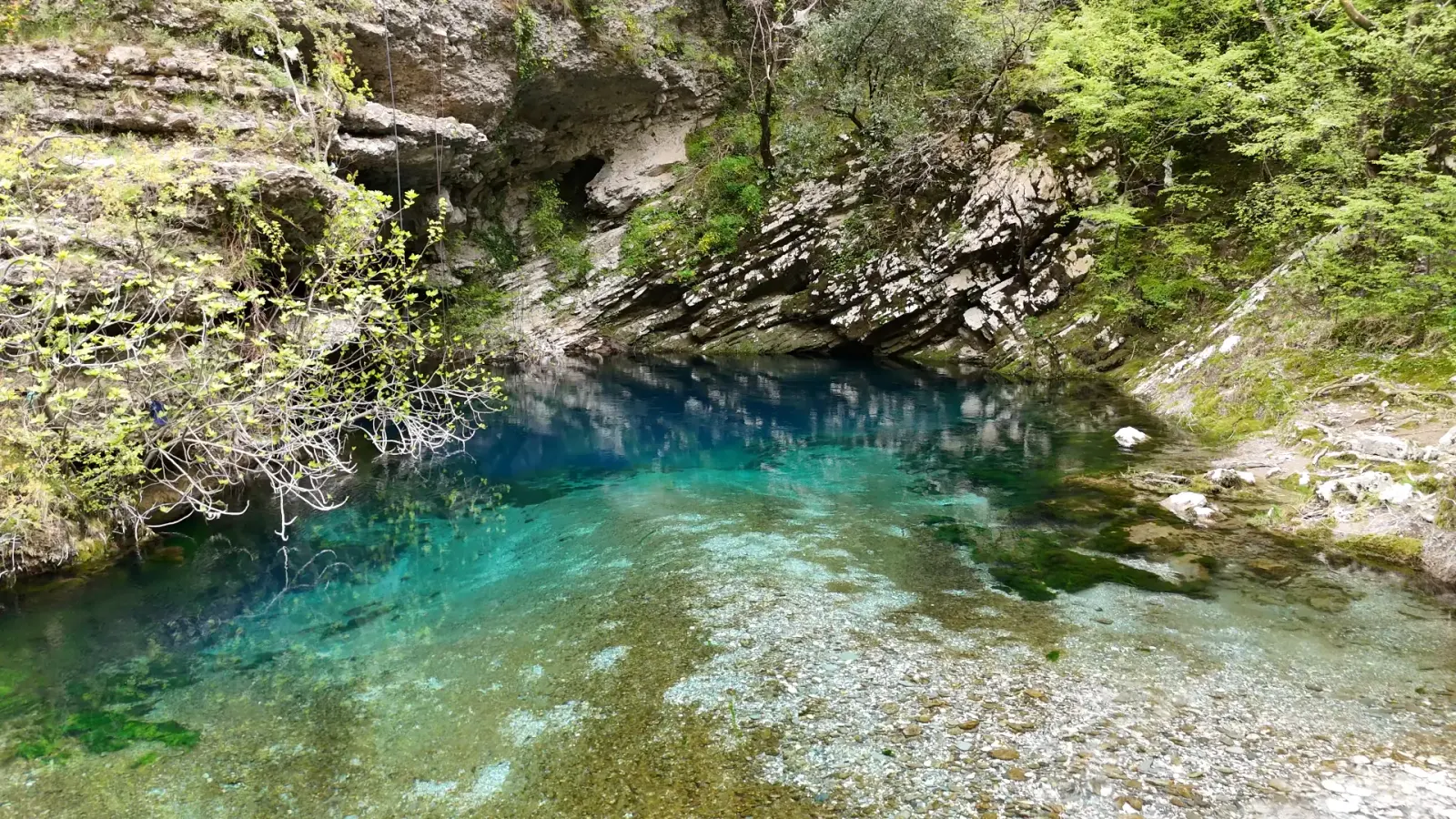 Clear turquoise pool of the Uji i Zi spring below a rocky overhang and greenery