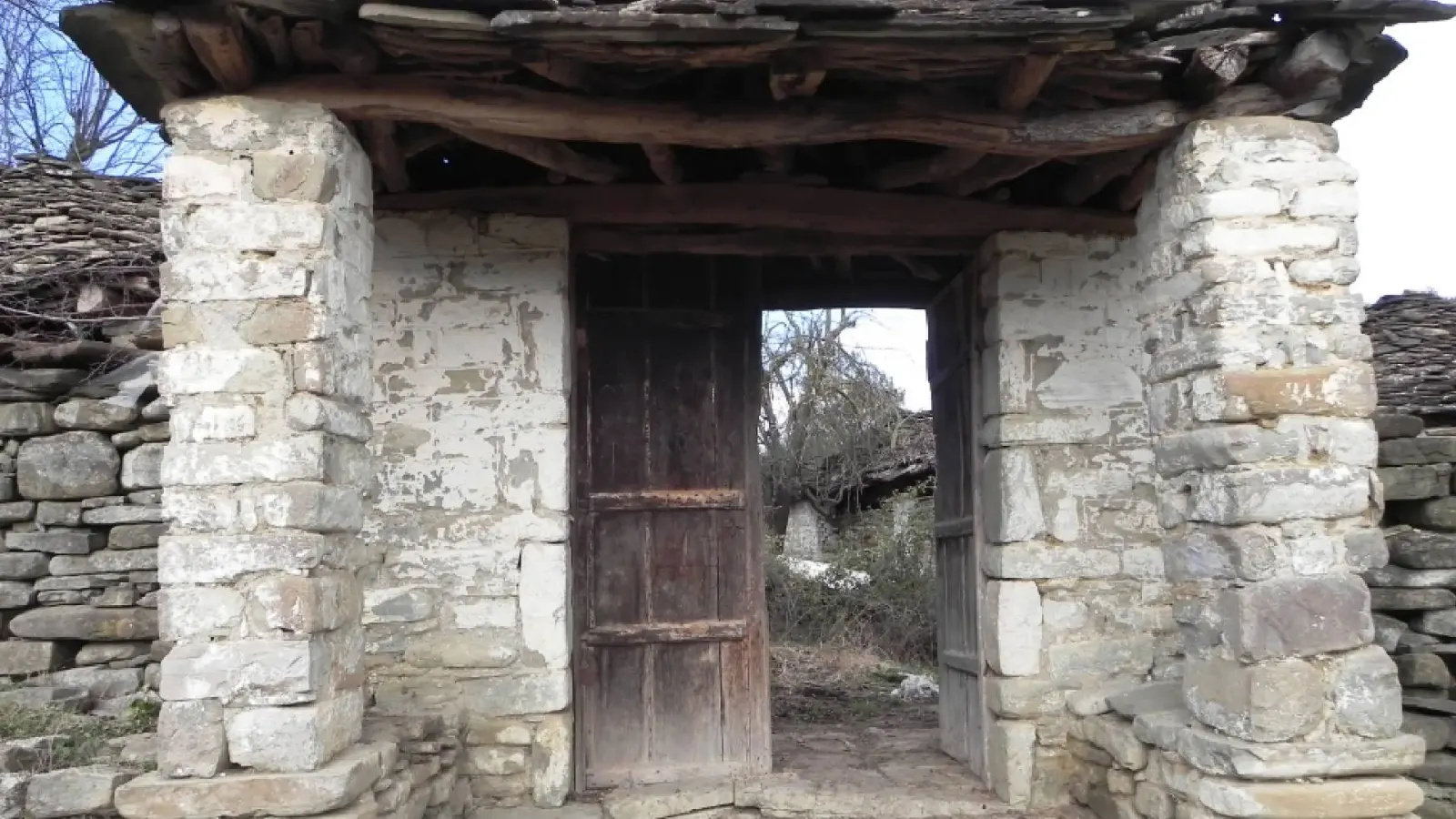 Überdachtes Steintor mit Holztür und Holzdach im Dorf Kenaj