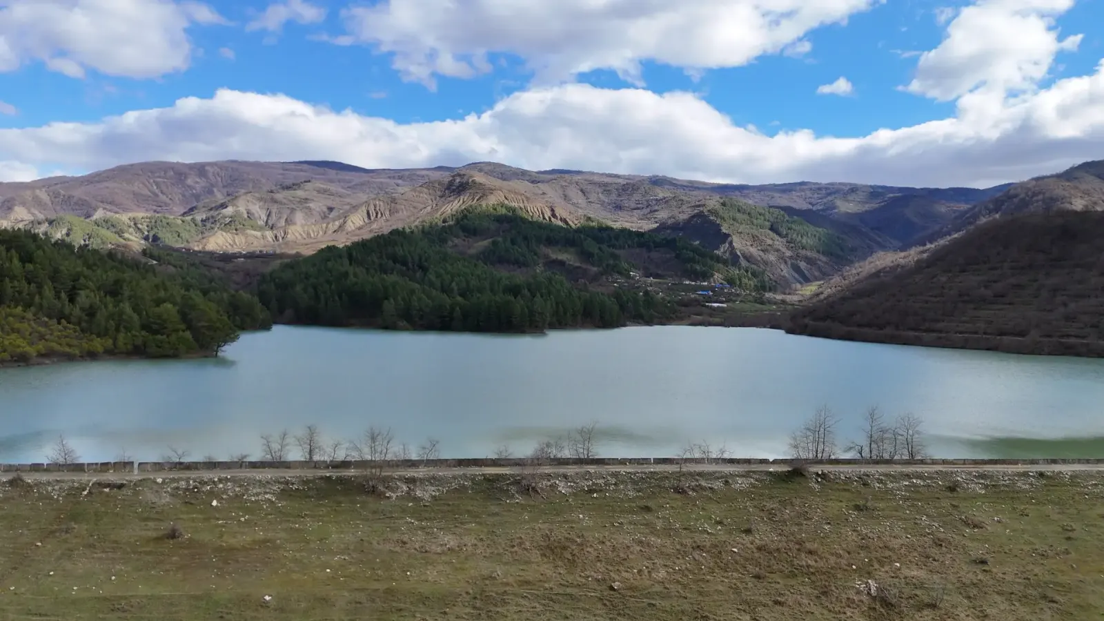 El embalse de Vinokash visto desde un terraplén de hierba con pinar y colinas al fondo